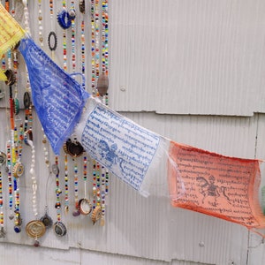 Puede incluir: Dos banderas de oraci&oacute;n con escritura tibetana cuelgan de una pared blanca. La bandera superior es azul con escritura blanca y la bandera inferior es naranja con escritura blanca. Las banderas est&aacute;n unidas a una cuerda de cuentas y tapas de botellas.