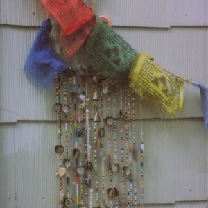 Puede incluir: Un carill&oacute;n de viento colorido hecho de cuentas, campanas y tapas de botellas cuelga de una pared de madera blanca. El carill&oacute;n de viento est&aacute; decorado con banderas de oraci&oacute;n coloridas.