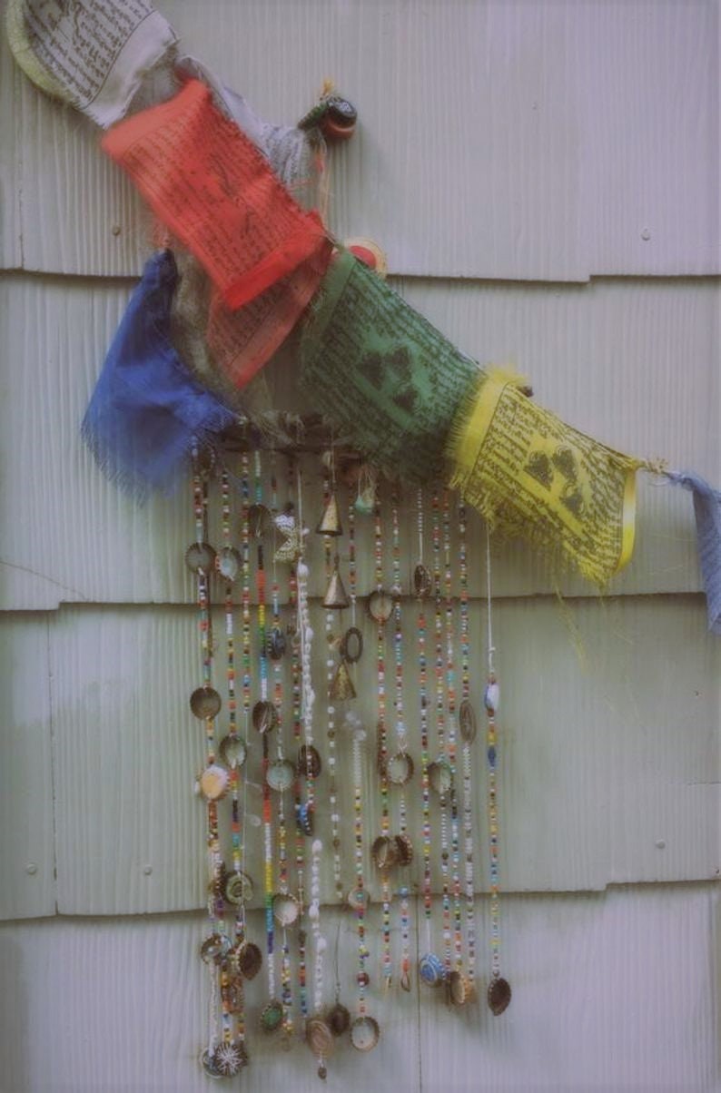 Puede incluir: Un carill&oacute;n de viento colorido hecho de cuentas, campanas y tapas de botellas cuelga de una pared de madera blanca. El carill&oacute;n de viento est&aacute; decorado con banderas de oraci&oacute;n coloridas.