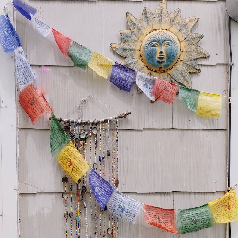 Puede incluir: Una pared blanca con una hilera de banderas de oraci&oacute;n coloridas y un sol de cer&aacute;mica con una cara azul. Un palo de madera cuelga de la pared con un carill&oacute;n de cuentas.