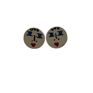 Puede incluir: Un par de pendientes redondos blancos con un diseño de cara pintado a mano. Cada pendiente presenta cabello negro, ojos azules, una nariz negra y una boca roja en forma de corazón.