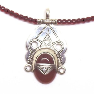 May include: A silver pendant necklace with a red oval stone and intricate etched designs. The pendant is suspended from a strand of small, round, red beads. The necklace is displayed against a white background.