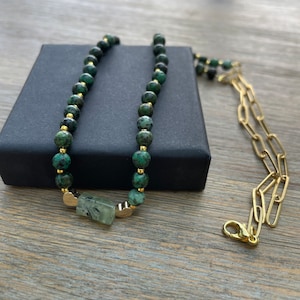 May include: A green gemstone necklace with gold accents and a heart-shaped pendant. The necklace is on a black box and a wooden surface.