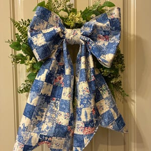 May include: A large decorative bow made of blue and white patchwork fabric featuring floral and toile patterns. The bow is attached to a green wreath with fern-like leaves and small white flowers, hanging against a paneled door.