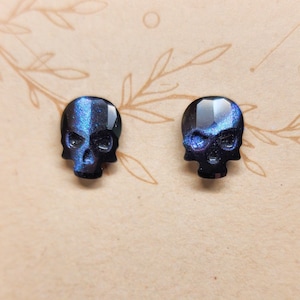 Peut inclure: Une paire de boucles d'oreilles clous en forme de crâne noir avec des accents de paillettes bleues irisées.