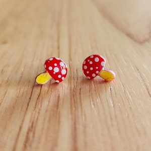 May include: A pair of red and white mushroom-shaped earrings with yellow stems. The earrings are on a wooden surface.