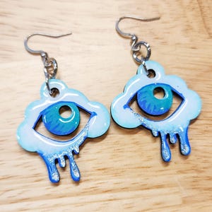 May include: A pair of unique earrings featuring a cloud and eye design. The earrings have a blue and white color scheme, with a stylized eye in the center of a cloud shape. The eye has a blue iris and a black pupil. The cloud has a dripping effect.