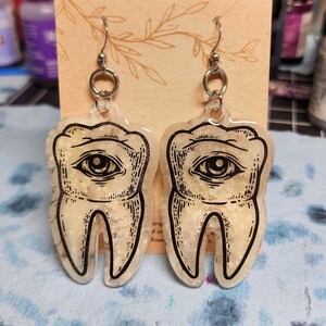 May include: A pair of silver earrings with a clear resin tooth design. Each tooth has a black eye with a white pupil and a white background. The teeth are translucent and have a speckled texture.