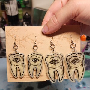 May include: A pair of silver earrings with a white tooth design featuring a black eye. The tooth is filled with glitter.