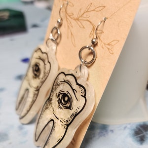 May include: A pair of silver dangle earrings with a clear resin tooth charm. The tooth charm has a black and white illustration of an eye on it.