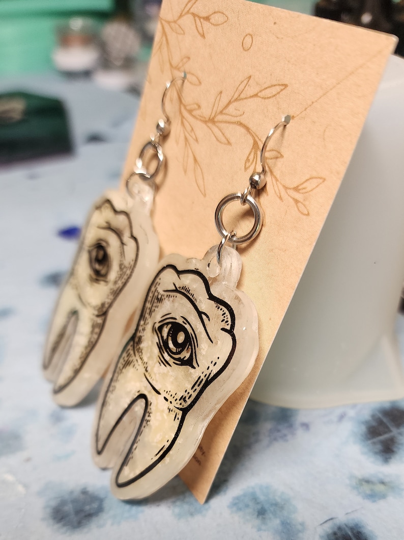 May include: A pair of silver dangle earrings with a clear resin tooth charm. The tooth charm has a black and white illustration of an eye on it.