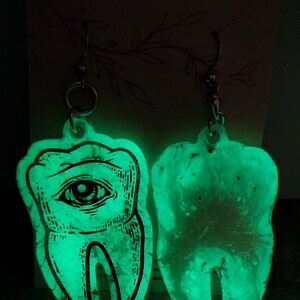 May include: A pair of glow-in-the-dark earrings shaped like teeth. One tooth has an eye design, while the other is plain. The earrings are made of clear resin with a glittery finish.