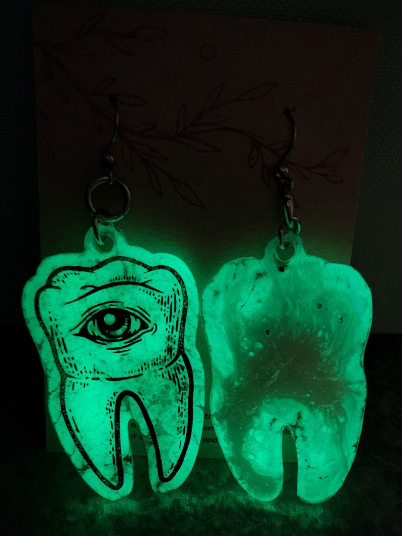 May include: A pair of glow-in-the-dark earrings shaped like teeth. One tooth has an eye design, while the other is plain. The earrings are made of clear resin with a glittery finish.