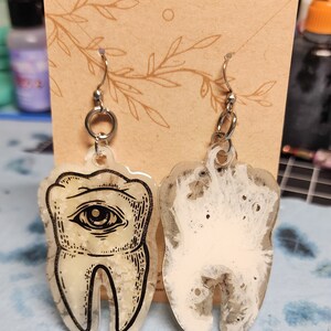 May include: A pair of resin earrings shaped like teeth. One earring has a black and white eye design, while the other is white with a translucent, cloudy effect.