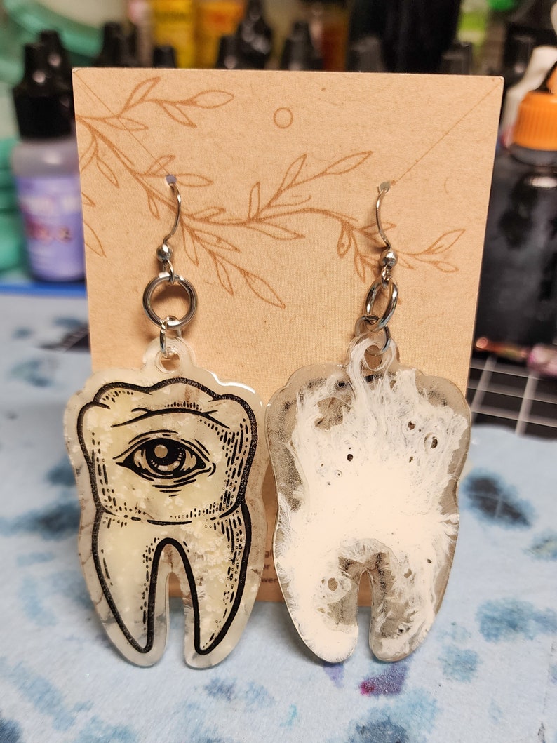 May include: A pair of resin earrings shaped like teeth. One earring has a black and white eye design, while the other is white with a translucent, cloudy effect.
