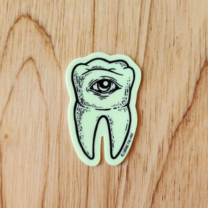 May include: A green glow-in-the-dark sticker of a tooth with an eye in the center. The sticker has the text "Alluring Collage" at the bottom.