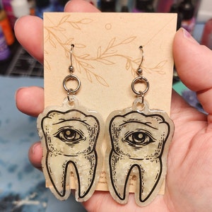May include: A pair of gold-tone earrings with a clear resin tooth design. Each tooth has a black and white eye illustration and a glittery texture.