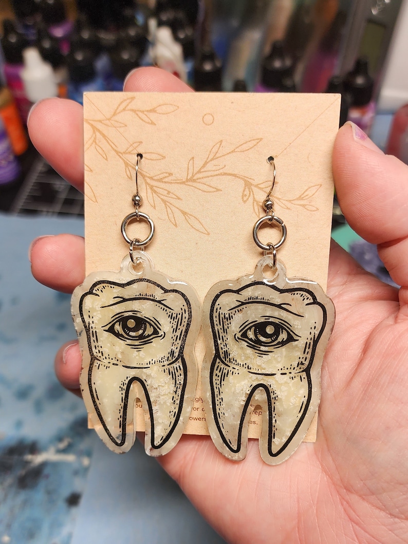 May include: A pair of gold-tone earrings with a clear resin tooth design. Each tooth has a black and white eye illustration and a glittery texture.
