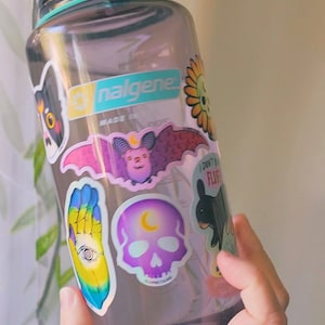 May include: A clear plastic water bottle with a teal lid and a gray base. The bottle is covered in colorful stickers, including a black cat, a bat, a skull, a hand with an eye, and a flower. The bottle is labeled "Nalgene" and "Made in China".