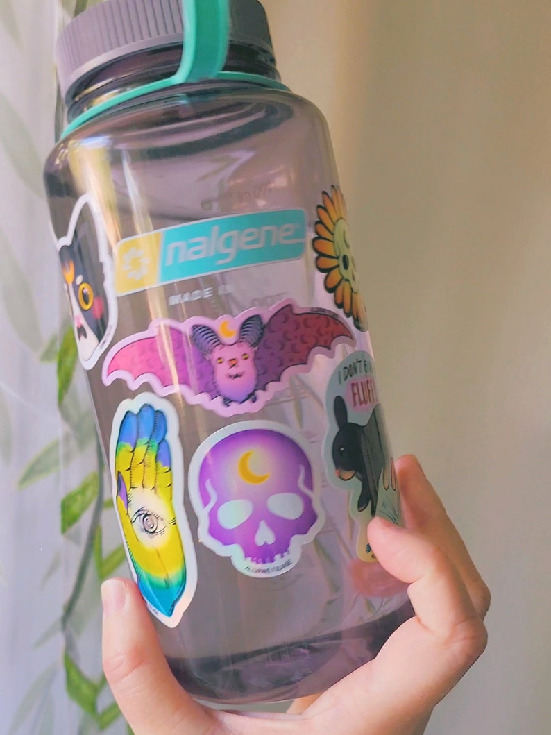 May include: A clear plastic water bottle with a teal lid and a gray base. The bottle is covered in colorful stickers, including a black cat, a bat, a skull, a hand with an eye, and a flower. The bottle is labeled "Nalgene" and "Made in China".
