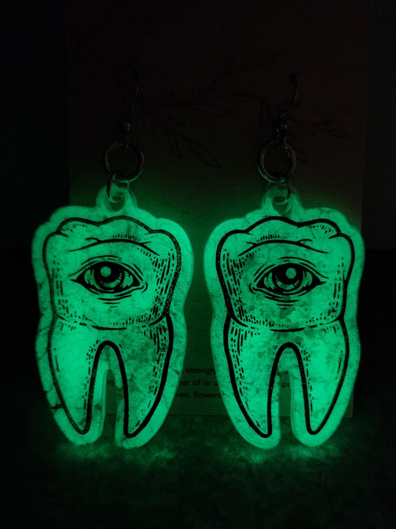 May include: A pair of glow-in-the-dark earrings shaped like teeth with an eye in the center. The earrings are made of clear resin with a glittery finish.