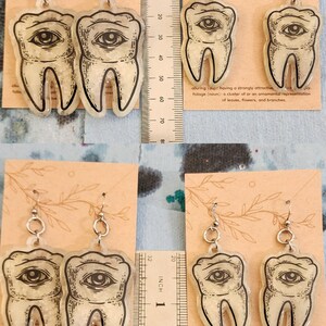 May include: A pair of silver earrings with a clear resin tooth design. The tooth has a black eye with a white pupil and is filled with glitter. The earrings are hanging from a ruler that is marked in inches.