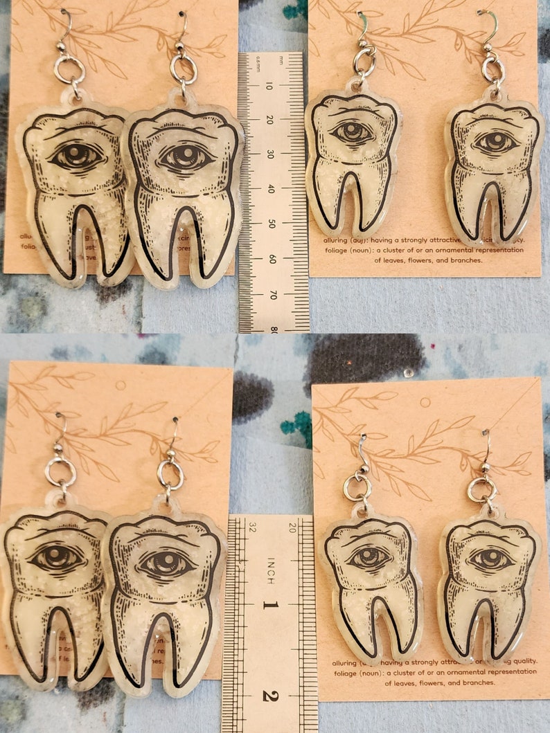 May include: A pair of silver earrings with a clear resin tooth design. The tooth has a black eye with a white pupil and is filled with glitter. The earrings are hanging from a ruler that is marked in inches.