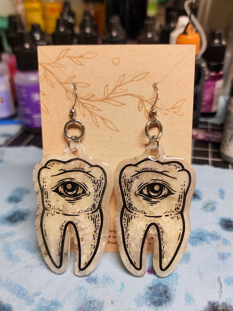 May include: A pair of silver earrings with a clear resin tooth design. Each tooth has a black eye with a white pupil and a white background. The teeth are translucent and have a speckled texture.