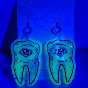 May include: A pair of glow-in-the-dark earrings shaped like teeth with an eye in the center. The earrings are made of clear resin with a black outline and are hanging from silver hooks.