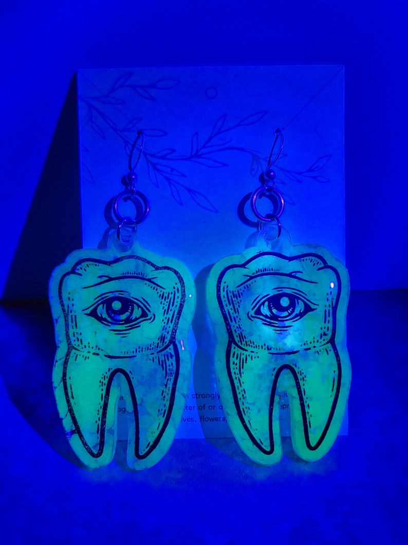 May include: A pair of glow-in-the-dark earrings shaped like teeth with an eye in the center. The earrings are made of clear resin with a black outline and are hanging from silver hooks.