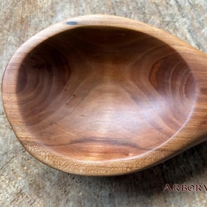 May include: A close-up of a wooden spoon with a deep bowl and a smooth, polished finish. The spoon is made of a light brown wood with a reddish hue. The grain of the wood is visible and adds to the rustic charm of the spoon.