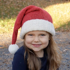 Op de afbeelding: Een kind draagt ​​een rood-witte gebreide kerstmanmuts met een pompon bovenop. De muts heeft een lange, puntige top. Het kind glimlacht en kijkt naar de camera. De afbeelding bevat de tekst "DROPS DESIGN" en "www.garnstudio.com".