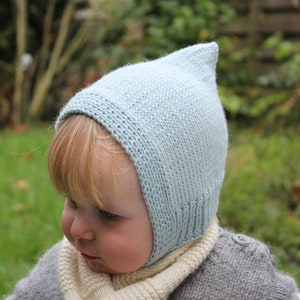 以下が含まれることがあります： 先のとがったトップを持つ薄い青色のニットのベビーボンネット。ボンネットはブロンドの髪の子供によって着用されています。