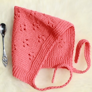 Op de afbeelding: Een roze gebreide baby muts met een strik. De muts heeft een kantpatroon en is gemaakt van zacht garen.
