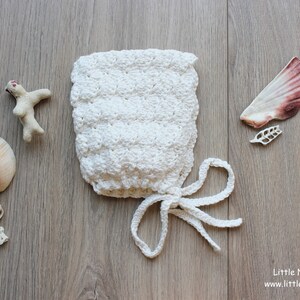 Peut inclure: Un bonnet crocheté blanc avec un motif en point coquille texturé et deux longs liens à la base. Le bonnet est entouré de divers coquillages et morceaux de corail. Le texte Little Natural Knits www.littlenaturalknits.nl est visible.