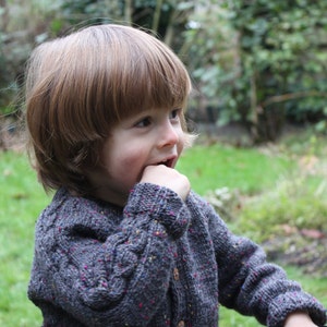 Könnte beinhalten: Ein kleines Kind mit braunen Haaren trägt einen grauen Pullover mit Zopfmuster und bunten Punkten. Das Kind beißt sich in den Finger und schaut nach rechts.