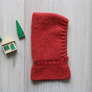 May include: A red knitted hooded scarf with a wood house and three green trees on a wood background.