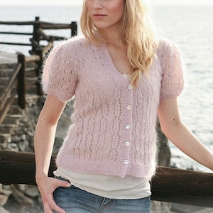 Peut inclure: Un cardigan en tricot rose clair à col en V et manches courtes. Le cardigan présente un motif de dentelle délicat et est boutonné sur le devant. Le mannequin porte le cardigan avec un jean et se tient près d'une clôture en bois.