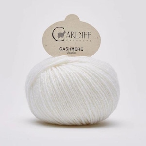 May include: A ball of white Cardiff Cashmere yarn with a label. The label is light brown with the brand name and a sheep logo. The text "CASHMERE classic" is printed on the label. The yarn is tightly wound and appears soft.