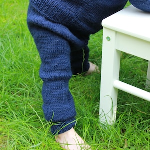 Puede incluir: Primer plano de polainas de punto azul para niños. Las polainas las lleva puestas un niño que está de pie sobre un taburete de madera blanco. Los pies del niño están descalzos y el taburete está en una zona cubierta de hierba.