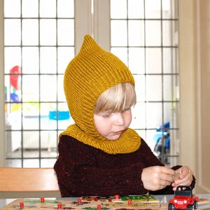 May include: A mustard yellow knitted bonnet with a pointed top and a ribbed texture that covers the head and neck, worn over a dark maroon textured sweater while playing with a small red toy vehicle on a wooden puzzle board.