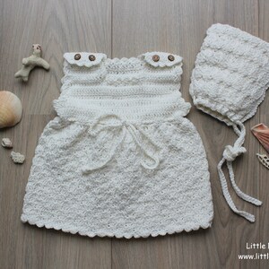 Puede incluir: Un vestido de ganchillo color crema con dos pequeños botones marrones en los hombros y un detalle de lazo en la cintura, acompañado de un gorro de ganchillo a juego. El texto Little Natural Knits www.littlenaturalknits.nl es visible en la esquina.