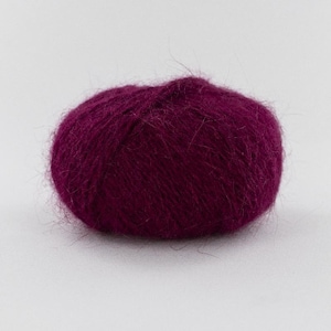 Puede incluir: Un ovillo de lana redondo de color magenta intenso con una textura suave, esponjosa y cepillada. Las fibras están densamente compactadas, creando una forma redondeada con un halo de hilos finos y sueltos que se extienden desde la superficie.