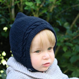 May include: A dark navy blue knitted bonnet featuring a pointed top and a textured cable knit border along the front edge. The bonnet is secured under the chin with a single visible button and is crafted from a soft, fine-gauge knit fabric.