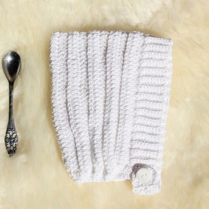 May include: A white crocheted baby bonnet with a button fastening. The bonnet is lying on a white fluffy surface.