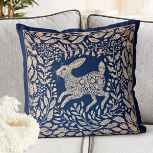 May include: A blue throw pillow with a white rabbit design. The rabbit is surrounded by floral patterns and leaves. The pillow is perfect for adding a touch of whimsy to your home decor.