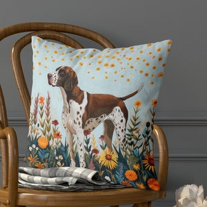 Op de afbeelding: Een kussen met een blauwe achtergrond waarop een bruine en witte hond staat in een veld met kleurrijke bloemen. De hond is een pointer met een lange snuit en slappe oren. De bloemen zijn in verschillende kleuren, waaronder geel, oranje, rood en paars.