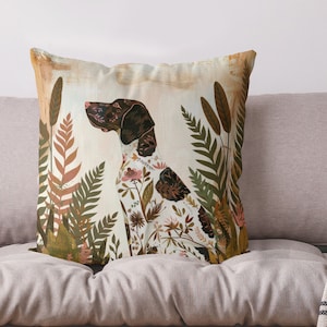 Könnte beinhalten: Ein Dekokissen mit einem braunen und weißen Hund, der in einem Feld mit Blumen und Farnen sitzt. Der Hund ist von grünen, braunen und rosa Blumen umgeben. Der Hintergrund ist hellbeige.