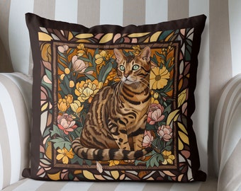 Peut inclure: Un coussin marron avec une bordure marron foncé présente un motif de vitrail représentant un chat tabby brun assis dans un lit de fleurs jaunes, orange et roses.