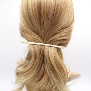 May include: A long, slender, gold-toned metal hair barrette with a smooth, slightly curved finish. The hair accessory is positioned horizontally across the back of a head of light blonde hair, securing a section of the hair in place.